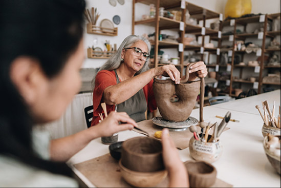classes-homepage Two women sculpting clay pottery in a ceramics studio, using tools and techniques for shaping and decorating.