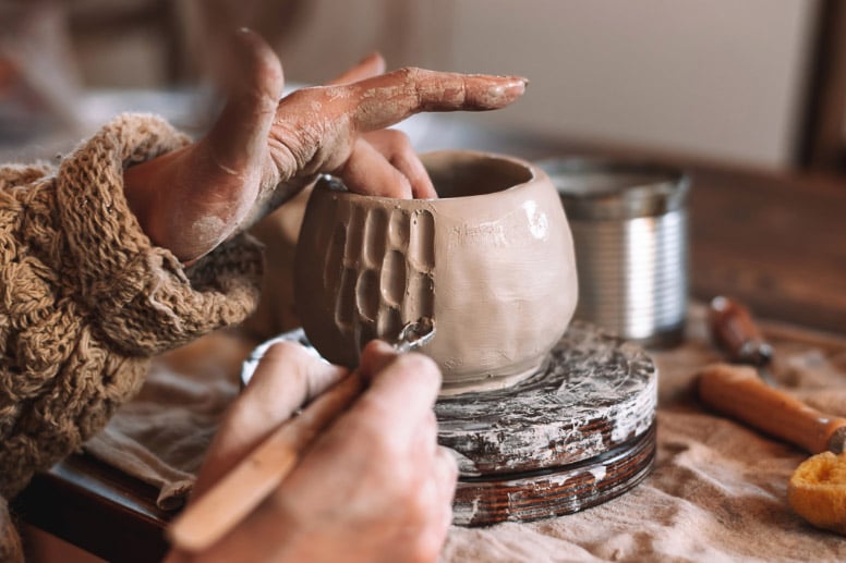 A person shaping clay on a pottery wheel, adding texture to a beige ceramic vessel with a wooden tool.