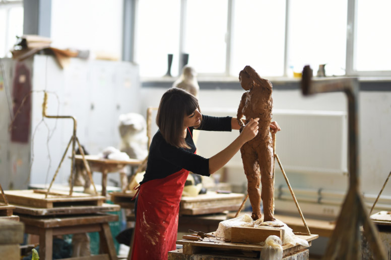 Artist shaping a clay sculpture in a ceramic studio, surrounded by tools, models, and kiln equipment.