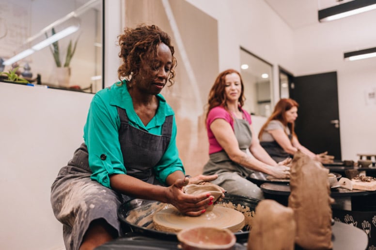 Three women working on pottery wheels, shaping clay while dressed in aprons, in a bright ceramics studio.