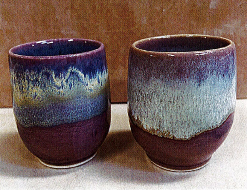 Two pottery mugs with rich purple, blue, and green glazes showcasing a gradation effect, set against a neutral background.