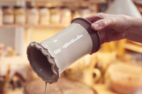 dipping-glaze A hand holds a scalloped-edge ceramic vase with glossy white glaze, dripping wet clay, in a pottery studio setting.