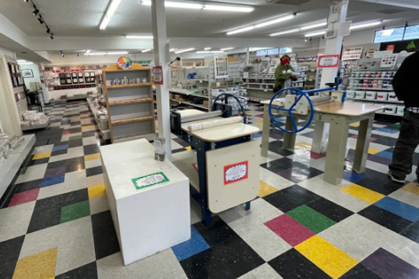 georgies-location-1 Brightly lit ceramics studio with rolling pin equipment, colorful tiled floor, and shelves displaying various pottery supplies.