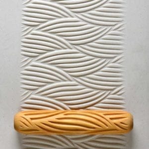 Textured clay mat showing wave patterns in white clay, with an orange rubber roller for embossing designs in ceramics.