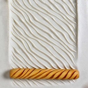 Texture roller with wavy pattern resting on textured white clay slab, used for decorating pottery and ceramics.