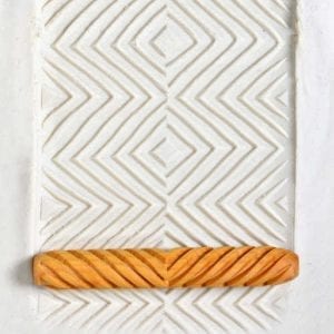 Patterned clay surface with textured zigzag impressions and a carved orange clay roller tool for ceramic decorating.
