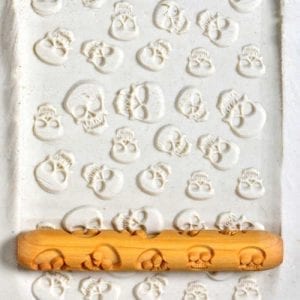 A white clay slab imprinted with skull patterns, complemented by a textured rolling pin with a skull design.