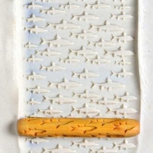 Impression rolled into clay featuring fish patterns, with a wooden brayer used for texture in ceramics.