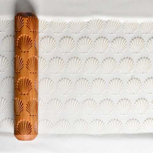 A textured rolling pin with shell designs beside an embossed clay surface featuring repetitive shell patterns.