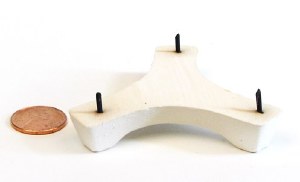White ceramic trivet with three metal spikes next to a penny for size reference, used for cooling pottery.