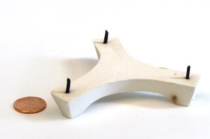 White ceramic shelf support with two black pins, alongside a penny for scale, used in kiln firing for pottery.