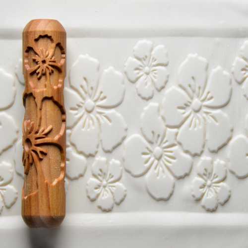 Wooden texture roller with floral design next to embossed white clay surface, ideal for pottery decoration.