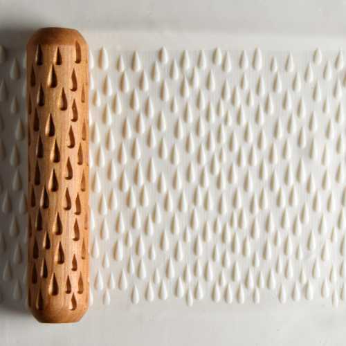 Textured pottery roller pressing into smooth white clay, creating a pattern of teardrop shapes for decorative ceramics.