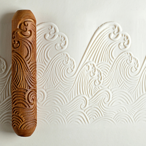 Wooden wave-patterned rolling pin beside embossed clay sheet, showcasing decorative techniques for ceramics and pottery projects.