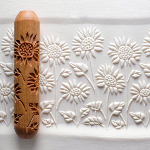 A carved wooden roller with sunflower designs resting on textured white clay, ideal for creating decorative pottery.