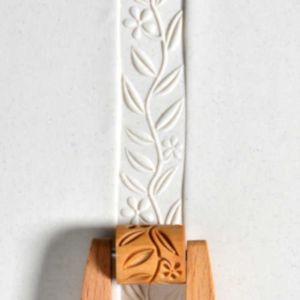 Impressed white clay ribbon with floral design and wooden decorative roller, displayed on a wooden stand for pottery techniques.