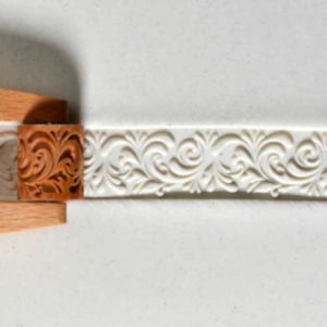 A wooden texture stamp presses a detailed, ornate pattern into soft white clay, showcasing intricate designs for pottery decoration.