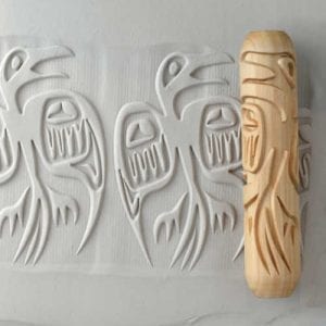 A wooden clay texture roller featuring intricate carved bird designs rests beside an impressed pattern on white clay.