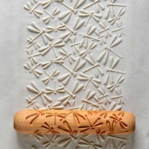 Wooden rolling pin with dragonfly imprint on white clay surface, showcasing texture for decorative pottery techniques.