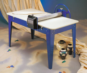 Blue slab roller with white surface, used for pottery preparation, beside decorated ceramic vases on a textured backdrop.