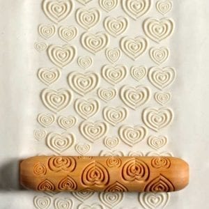 A wooden rolling pin presses into soft clay, creating a pattern of embossed hearts on the surface.