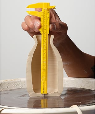 A hand holds a caliper measuring the height of a wet clay vase on a pottery wheel, showcasing ceramic techniques.
