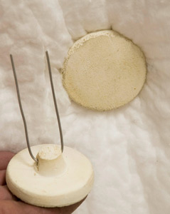 A hand holding a ceramic clay base with wire supports, next to a textured clay piece on a soft surface.