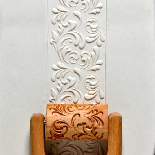 Embossed white clay adorned with a swirling design, pressed by a carved wooden roller for decorative pottery techniques.