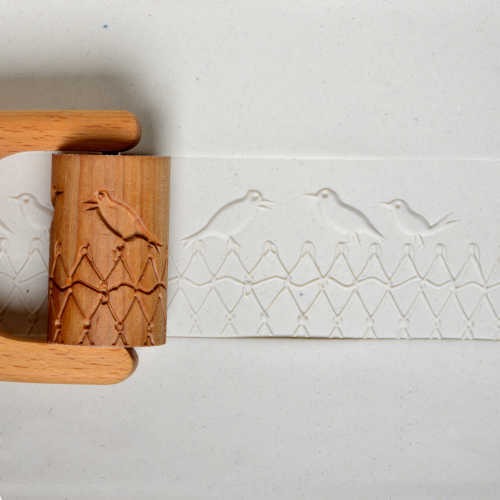 Wooden texture roller with bird designs next to an embossed clay strip, showcasing ceramic decoration techniques.