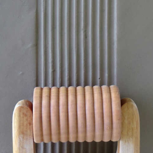 Wooden rib tool creating parallel grooves in a slab of gray clay, demonstrating texturing techniques for pottery.