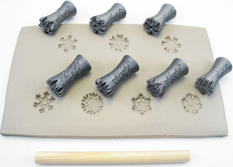 Six textured ceramic stamps on a slab of gray clay, featuring snowflake impressions and a wooden roller in the foreground.