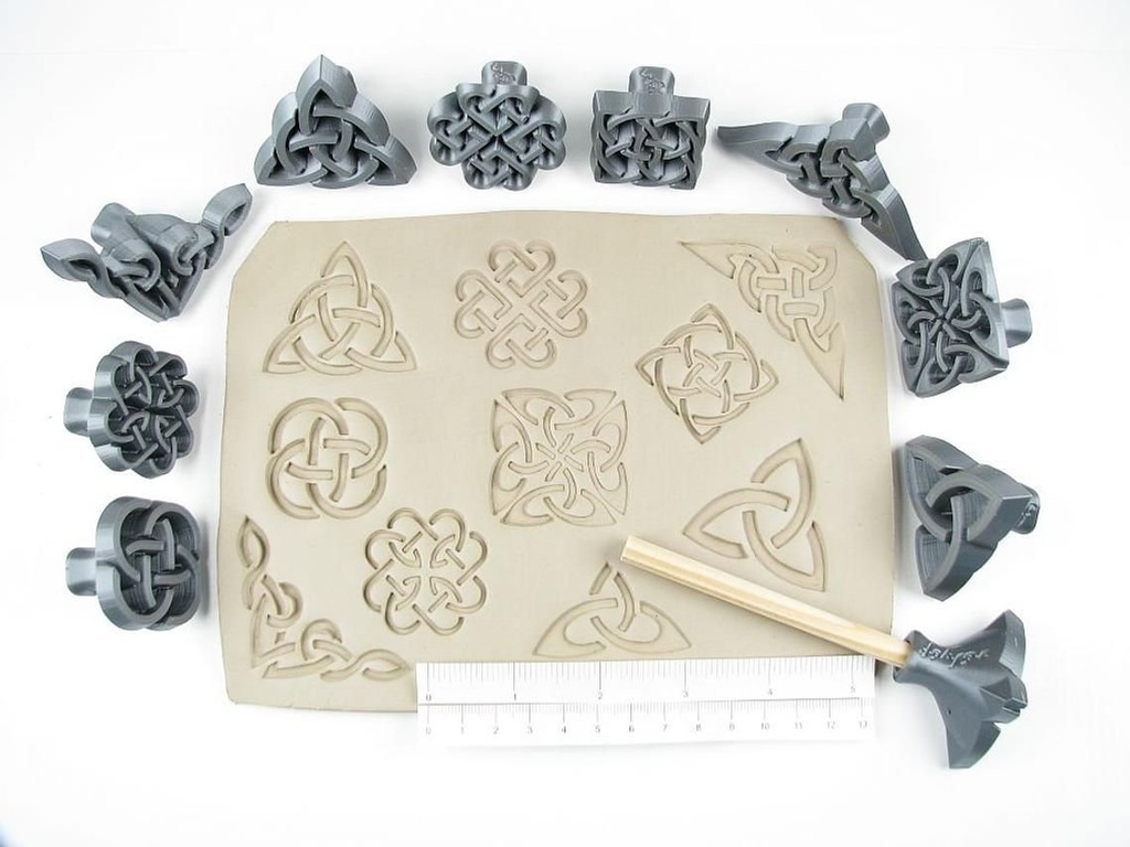 Various grey clay stamps arranged around an untrimmed slab of clay with intricate Celtic designs. A wooden tool and ruler are included.