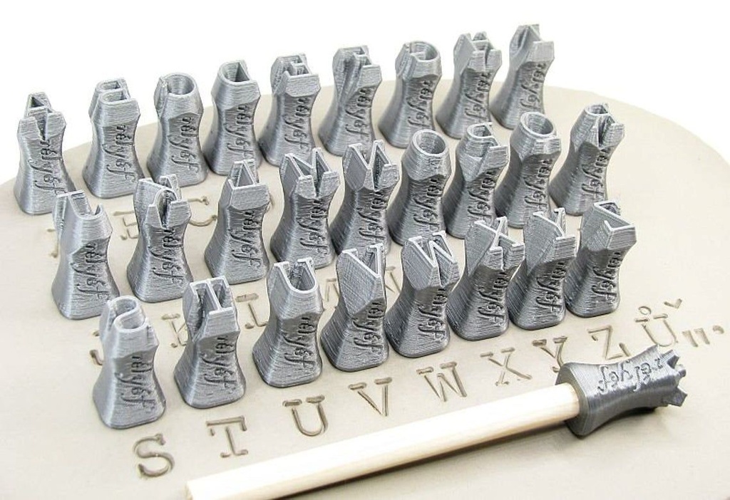 Twenty-eight small, gray clay letter stamps arranged on a clay slab, next to a stamping tool for decorating pottery.