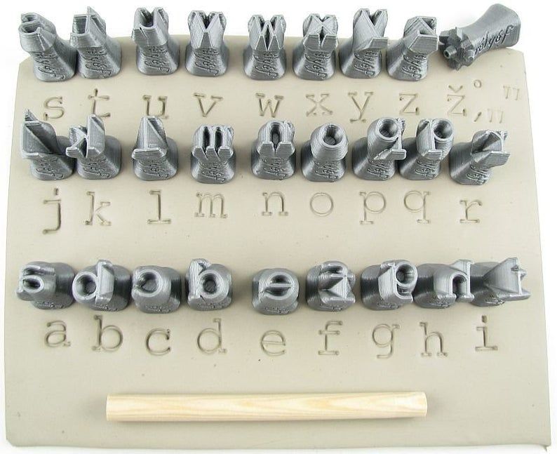 Ceramic alphabet stamps arranged on a slab of clay, with a wooden handling tool for pressing letters into pottery.