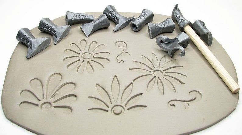 Clay slab with decorative tools and a wooden sculpting stick, featuring stamped floral patterns in the damp clay.