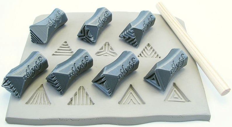 Gray clay stamps for texturing, arranged on a slab of soft clay with triangle impressions, alongside a wooden modeling tool.