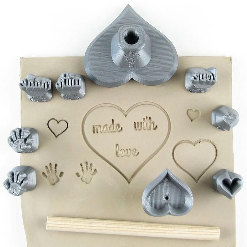 Grey clay stamps and tools arranged on a sheet of beige clay, featuring impressions of hearts and handprints, labeled made with love.