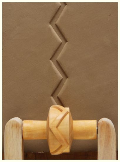 Wooden texture roller with zigzag pattern imprints on a slab of brown clay, showcasing pottery decorating techniques.