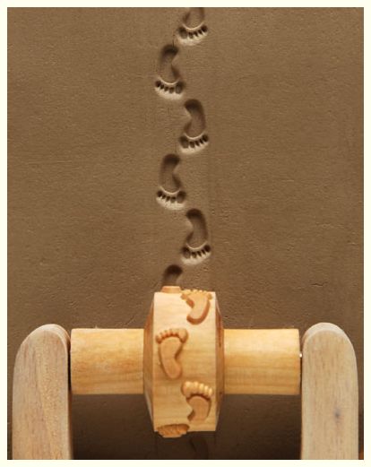Textured clay showing a rolling stamp with footprint design, creating impressions in the soft surface for pottery decorating.