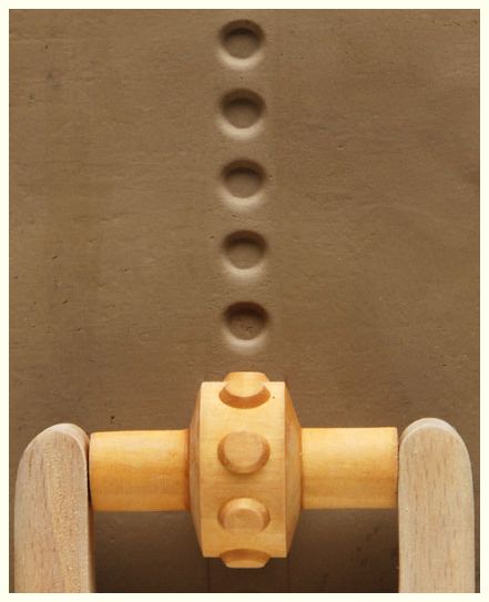 A close-up of a wooden roller tool creating evenly spaced circular impressions in smooth clay, ready for pottery shaping.