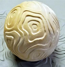 Textured clay sphere with engraved undulating patterns, displayed on a clay surface, showcasing pottery design techniques.
