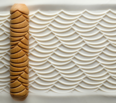 Wooden texture roller creating a wave pattern on smooth white clay, showcasing the technique for decorating pottery.