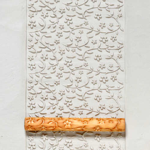 Embossed white clay slab featuring a floral pattern, with a textured wooden rolling pin used for imprinting designs.