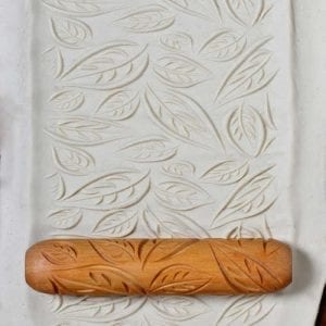 Embossed clay sheet featuring leaf patterns, with a wooden rolling pin used for texture creation in pottery.
