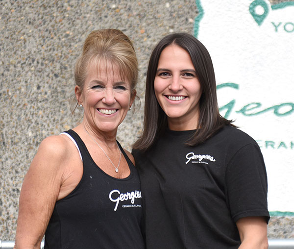 Two women smiling in front of a Georgies Ceramic & Clay Company sign, showcasing the community of ceramic enthusiasts.