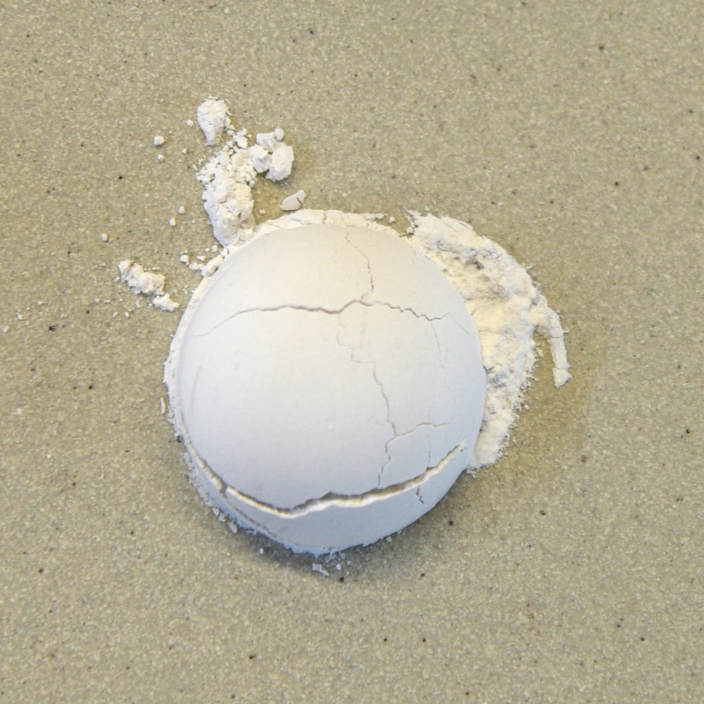 Cracked white ceramic sphere with powdery residue on a light-colored surface, likely from casting or molding clay.