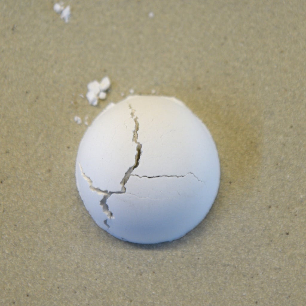 Cracked white ceramic ball resting on a sandy surface, illustrating potential defects in clay work or bisque firing.