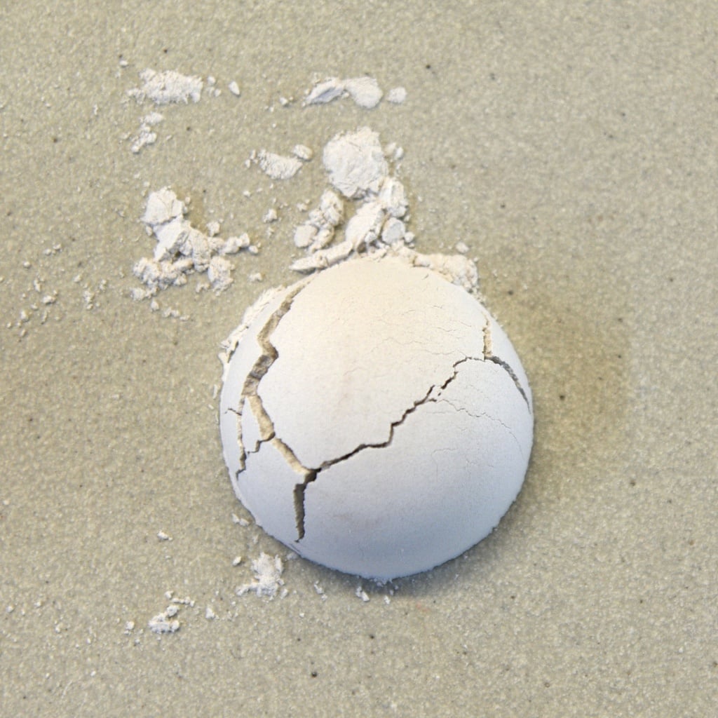 Cracked white ceramic ball on gray clay surface with scattered powder, showcasing texture and breaking under pressure.
