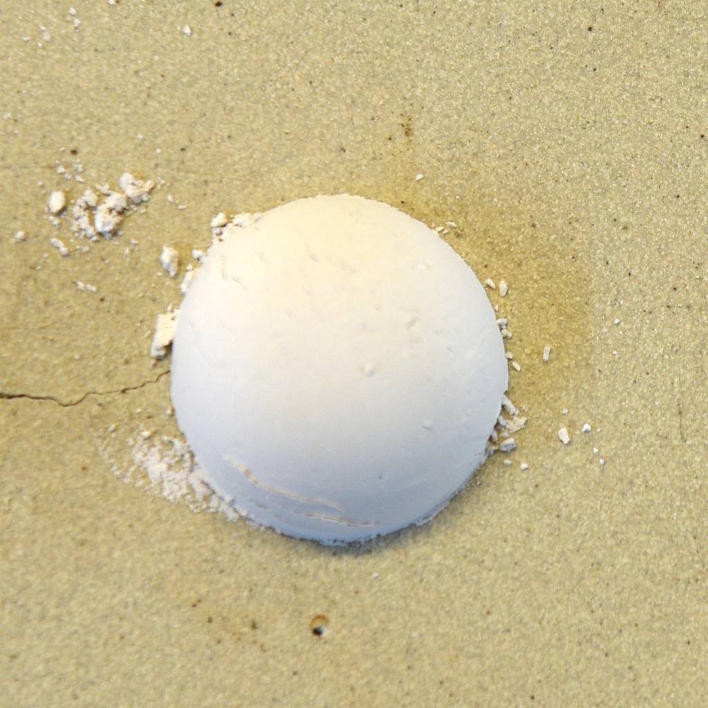 A smooth white ball of clay sits on a textured beige surface, surrounded by scattered dry clay particles.
