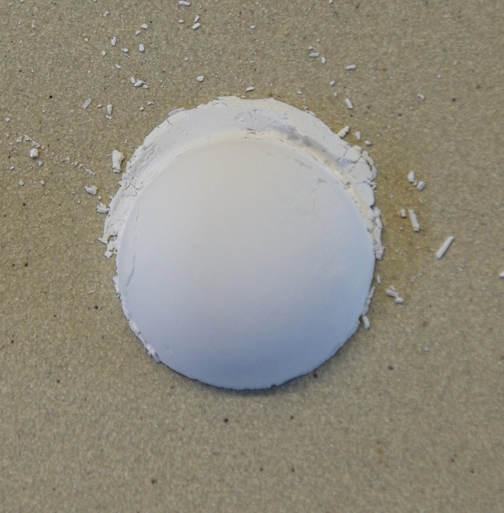 A smooth, white clay test piece sits on a sandy surface, with small clay shavings scattered around it.